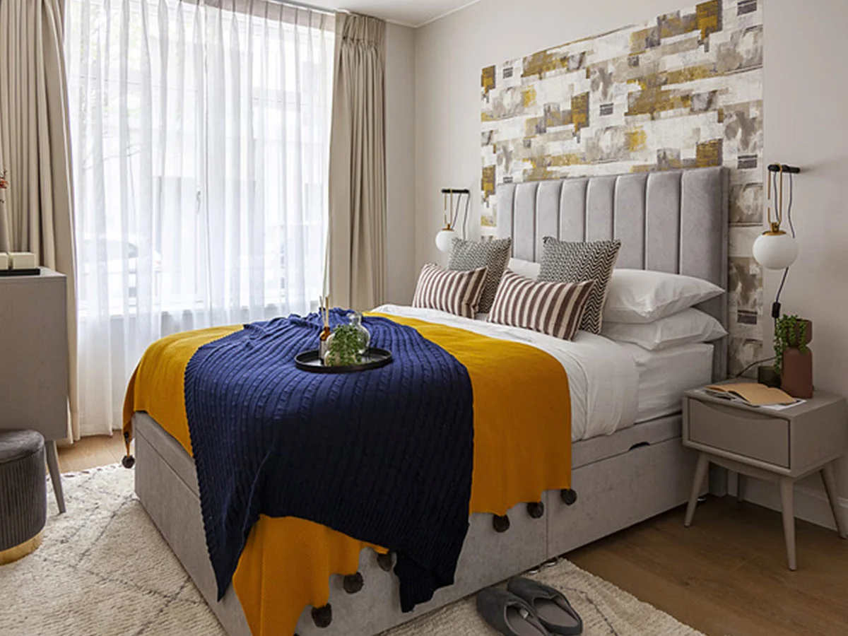 Modern bedroom with grey upholstered bed, mustard and navy throw, and patterned accent wall