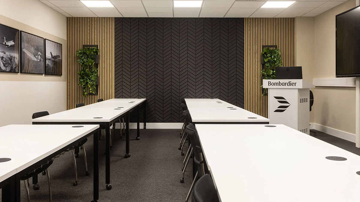A professional corporate training room styled by InStyle Direct featuring rows of white workstations, a black herringbone feature wall with vertical wood slats, integrated greenery, and a branded Bombardier lectern.