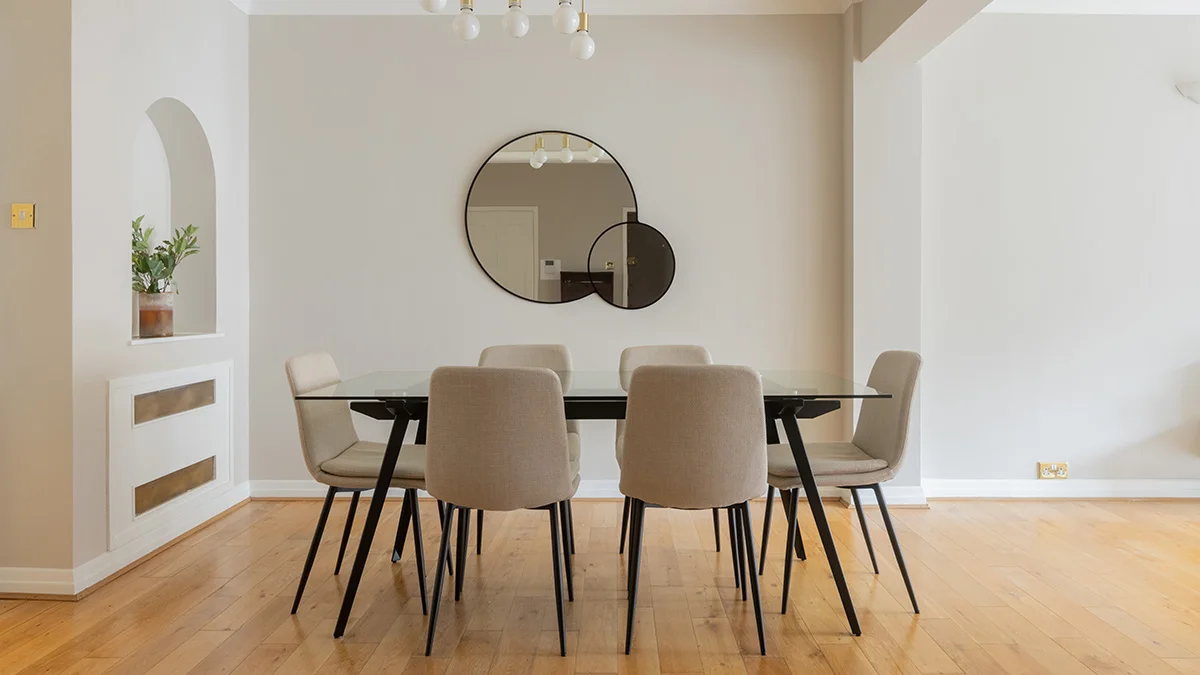 A professionally staged dining room by InStyle Direct featuring a sleek black-framed glass dining table with six neutral upholstered chairs, a contemporary double-circle wall mirror, and light oak wood flooring in London.