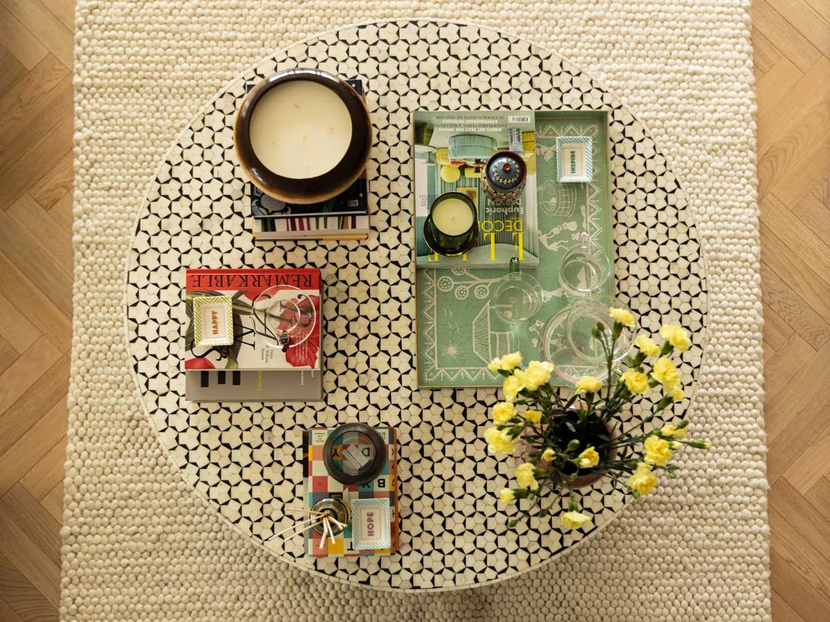 A bright living space featuring a minimalist black metal X-frame console table styled with designer books and a tall green lamp, positioned next to floor-to-ceiling sheer white curtains in a Riverside apartment