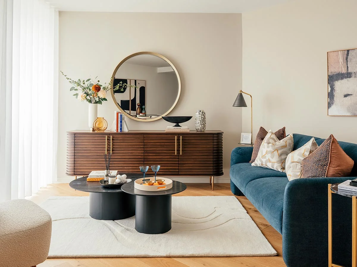 A professionally styled living room designed to help estate agents market properties, featuring an elegant blue sofa, a slatted wood console, and a large round mirror