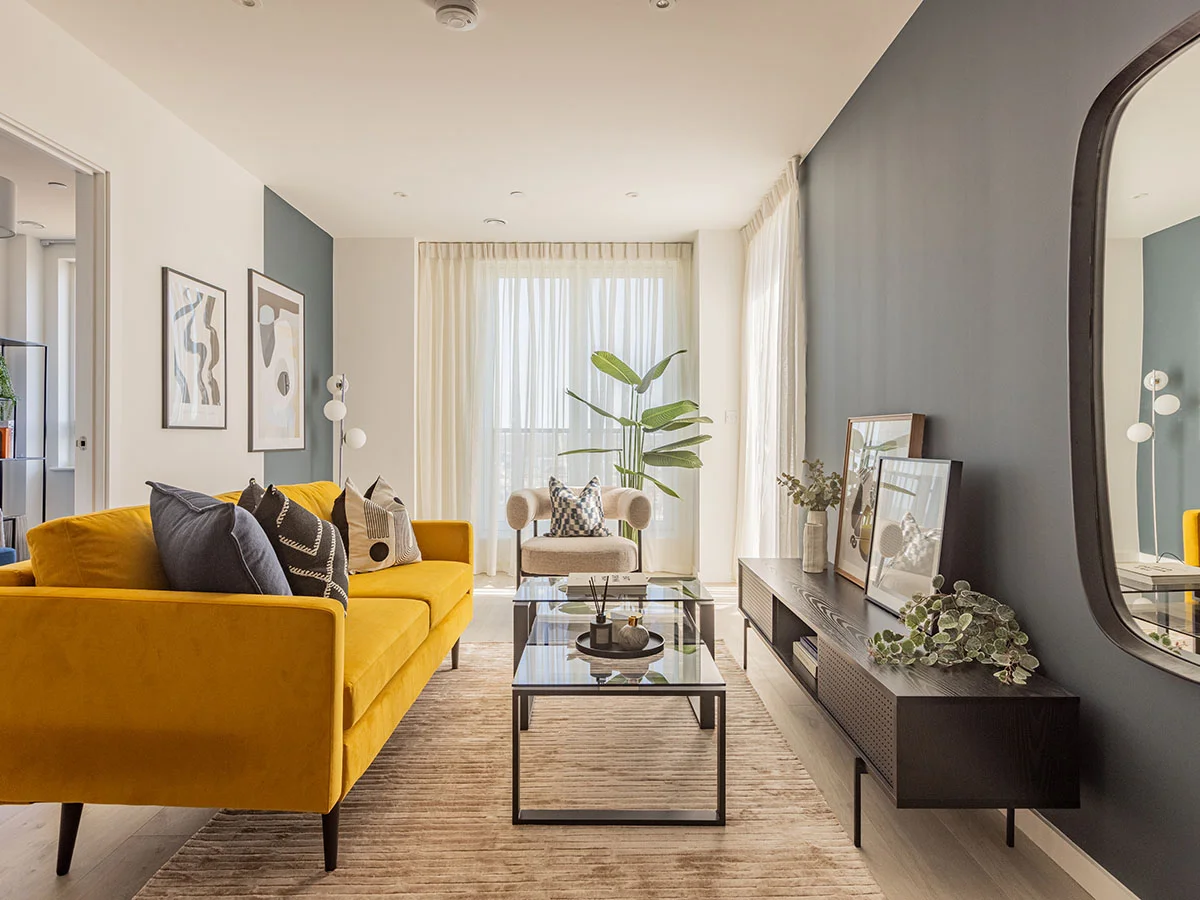 A vibrant living room professionally styled by InStyle Direct featuring a mustard yellow velvet sofa, a teal feature wall, glass-topped coffee tables, and a large black-framed mirror reflecting a modern apartment in London.