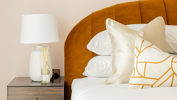 Bedroom corner with a mustard headboard, white bedding, and a bedside table with a lamp and diffuser, showcasing turnkey furnishing Dubai style