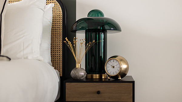 Stylish bedside setup with white bedding, a green glass lamp, gold clock, and diffuser on a wooden nightstand, reflecting studio furniture Dubai aesthetics