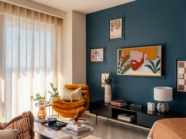 High-impact living room with navy accent wall, yellow chair, and framed TV art. Professional furnishing transformation appeal.